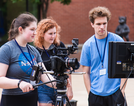 Three students film with a camera and watch the playback on a screen.